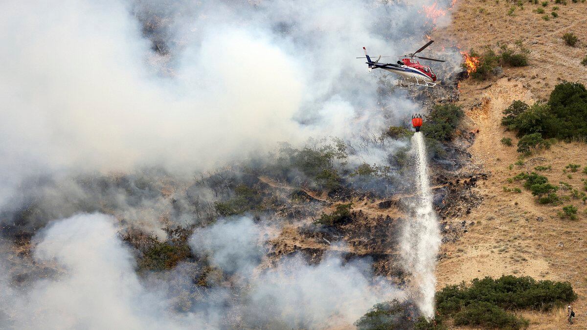 Firefighters battle a wildfire from the ground as a helicopter drops water above them in...
